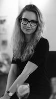 Portrait photo of Agne gently smiling and looking directly into camera. Agne has long wavy blonde hair, dark-rimmed glasses, and is wearing a black long-sleeved top. The photo is in black and white.