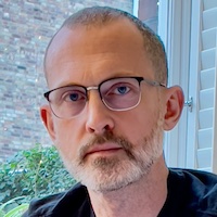 A man with short hair, a short beard and glasses in front of a window
